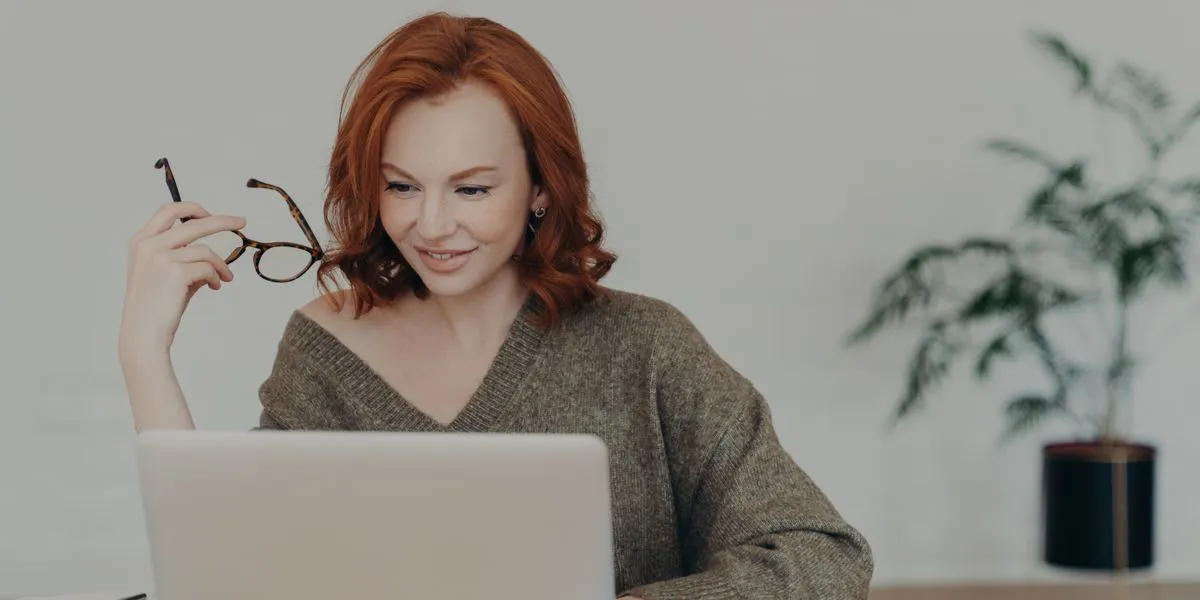 Mulher ruiva com os óculos na mão verifica no PC um trabalho Mulher ruiva com os óculos na mão verifica no PC um trabalho